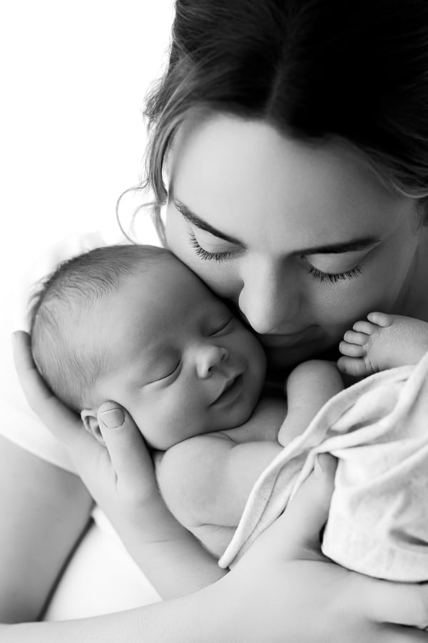 A mother kissing her sleeping newborn's head against a bright white background