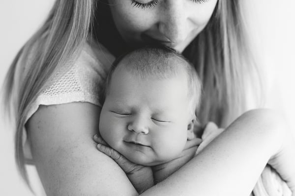 A mother holding her swaddled newborn against her chest and kissing the top of the baby's head, in black and white