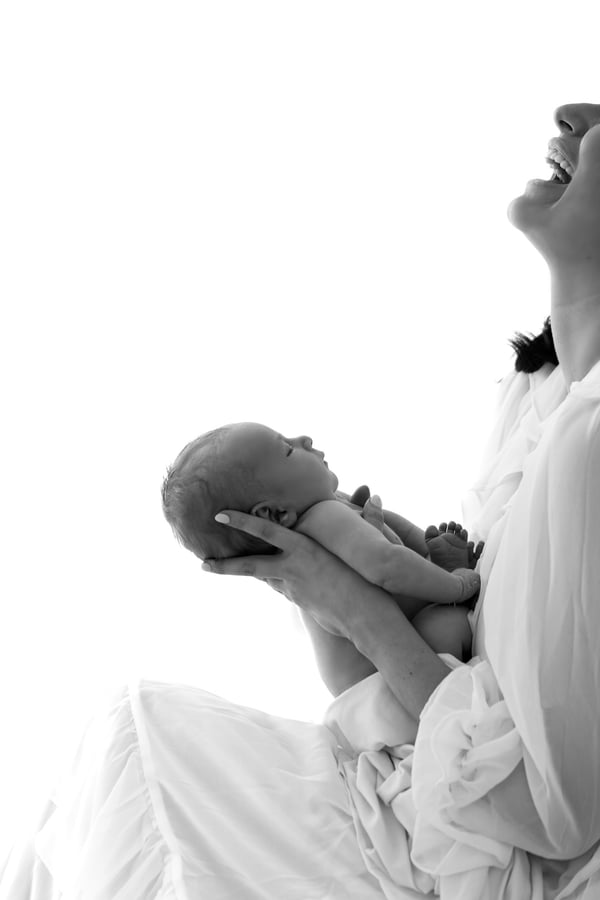 A mother laughing, head tipped back, lifting her newborn in both hands against a bright white background