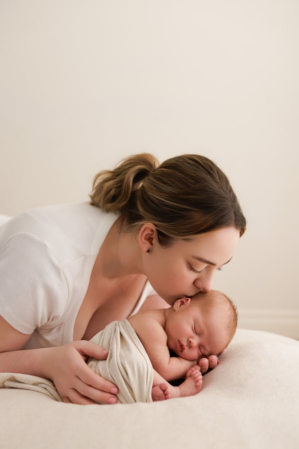 A mother leaning down to kiss her sleeping newborn, who lies wrapped on the beanbag