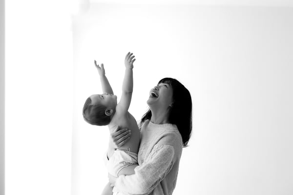 A mother laughing as she lifts her baby up above her against a bright window, in black and white