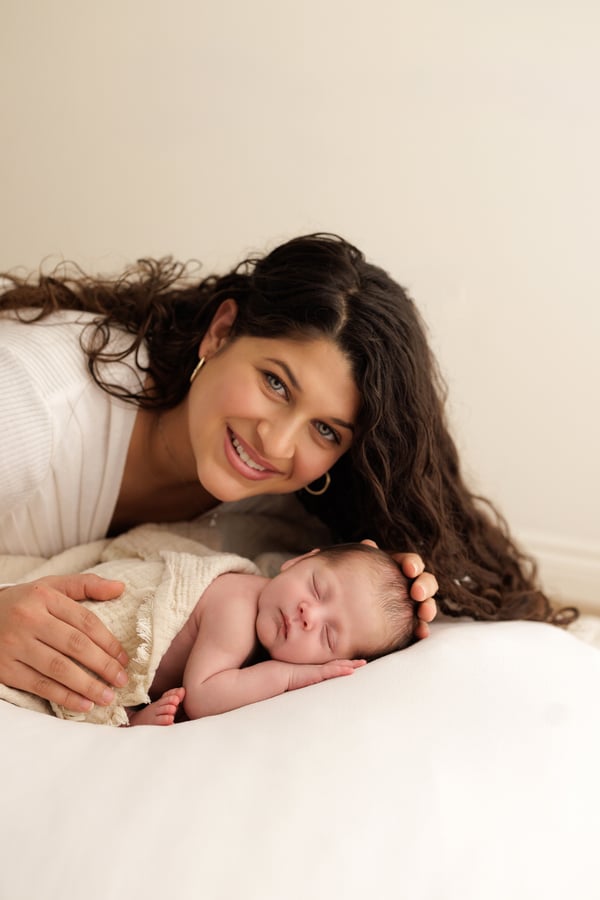A mother lying behind her sleeping newborn on a white blanket, smiling at the camera