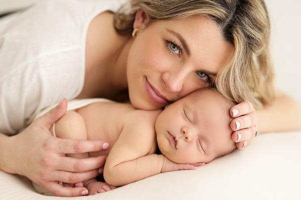 A mother lying beside her newborn with a hand resting on the baby, looking towards the camera