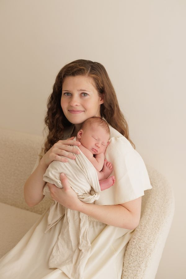 A mother seated in an armchair holding her swaddled newborn, looking at the camera