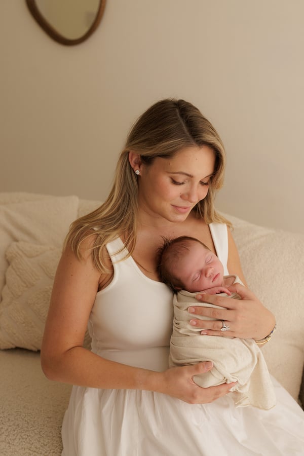 A mother seated in a pale armchair holding her swaddled newborn and looking down at her