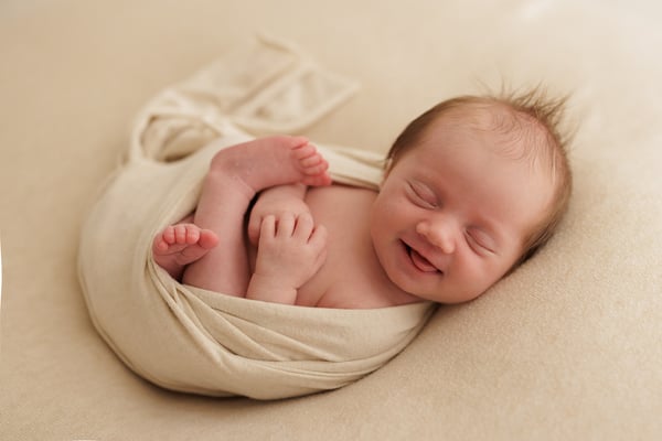 A newborn asleep in a cream wrap with the corners of her mouth turned up