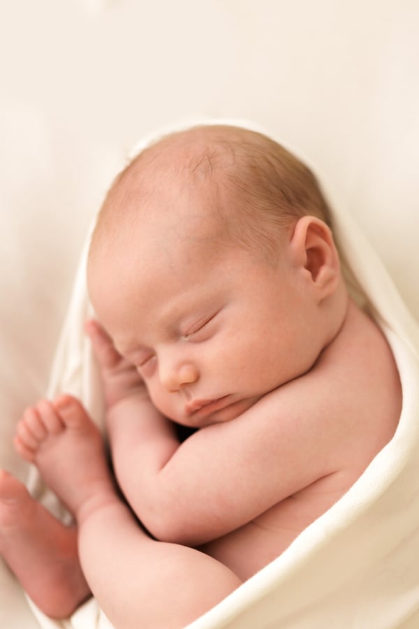 A newborn asleep on a cream wrap, curled with the feet tucked up
