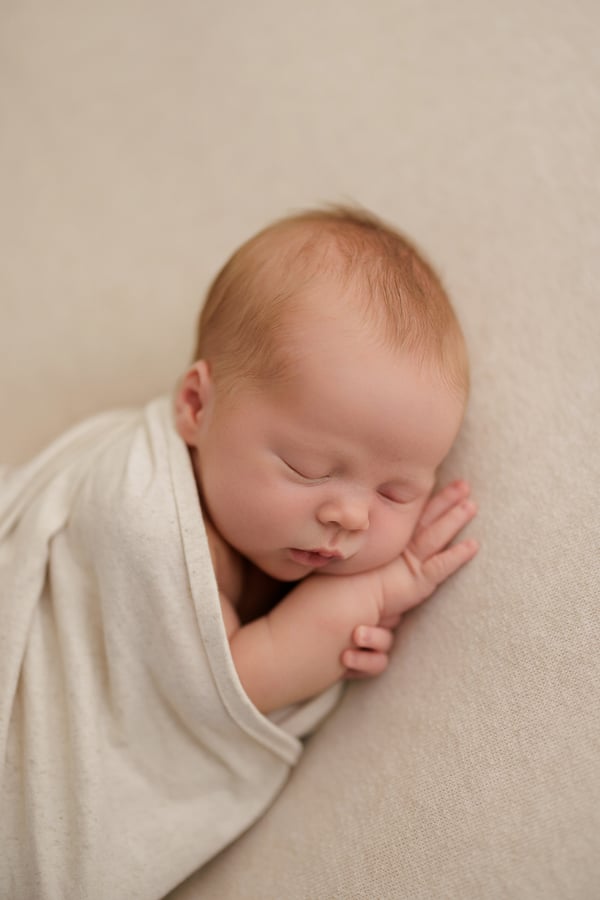 A newborn asleep in a cream wrap, seen from the side on a cream blanket