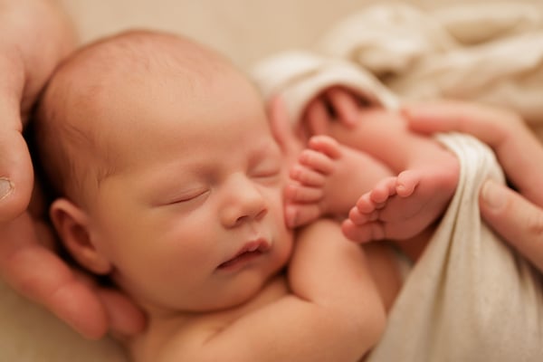 Newborn asleep cradled in two hands, tiny feet showing beyond the wrap