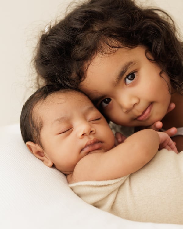 A big sister resting her cheek against the sleeping newborn beside her, looking at the camera