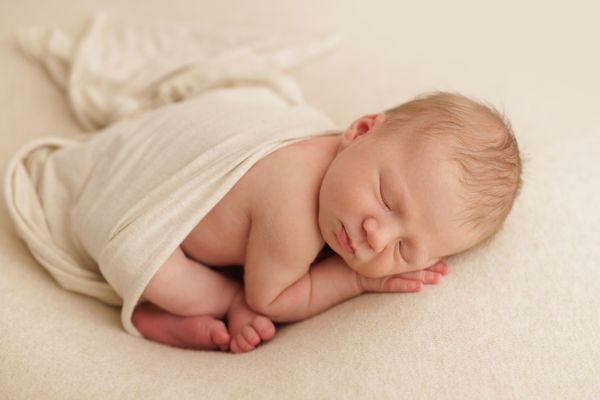 A newborn asleep in a cream wrap on a cream backdrop, feet showing