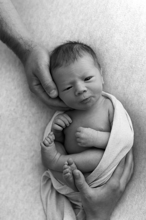 A newborn awake in a light wrap, a hand cradling their head on a textured blanket, in black and white