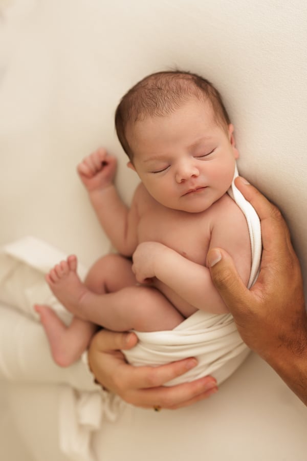 A newborn seen from above on a cream backdrop, one arm raised, a parent's hand supporting