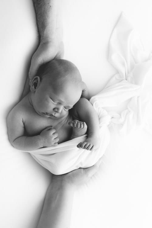 Newborn lying on its back on a white wrap, head resting in a parent's hand