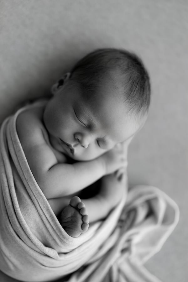 Newborn curled up in a wrap on a grey backdrop, one foot showing, in black and white