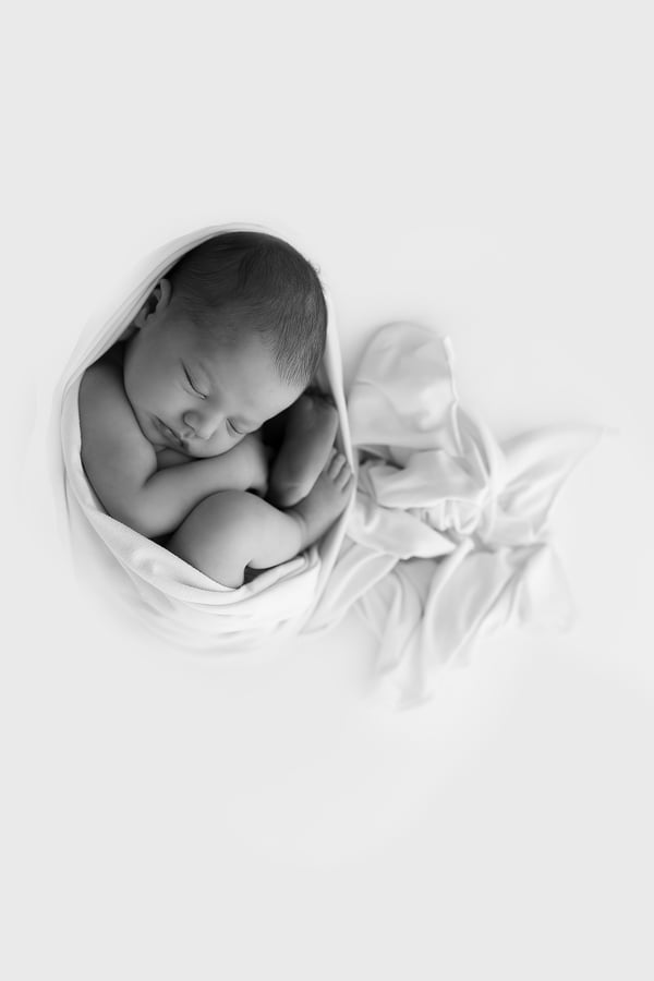 Newborn wrapped in white, small within a bright empty frame, in black and white