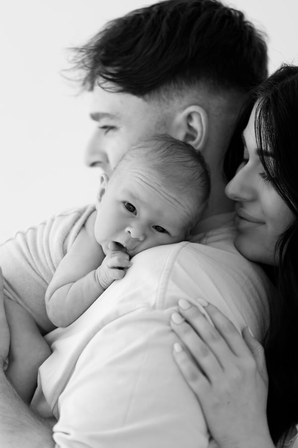 Both parents cheek to cheek, holding their newborn, who is awake and looking out from the wrap