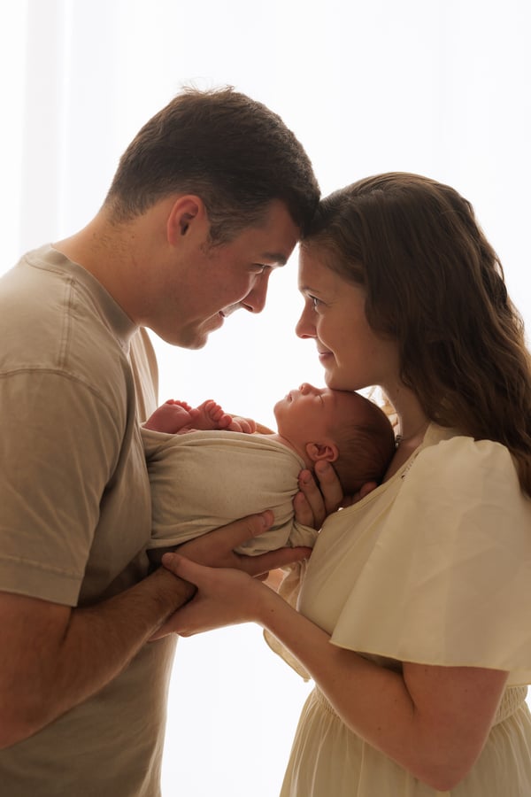 Both parents standing forehead to forehead, holding their swaddled newborn between them against a bright window