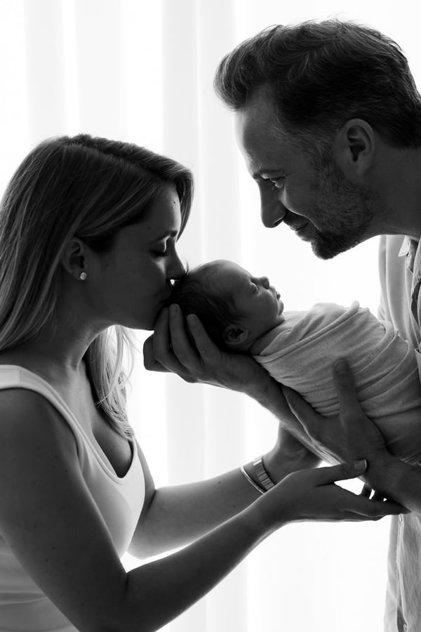 Both parents holding their newborn up between them against a bright window, in black and white
