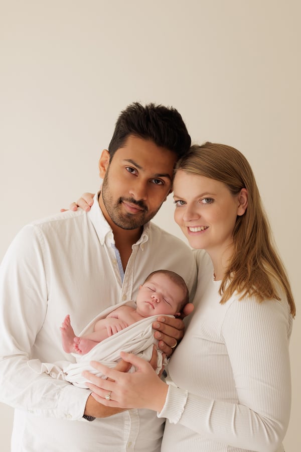 Both parents in white holding their swaddled newborn and looking at the camera