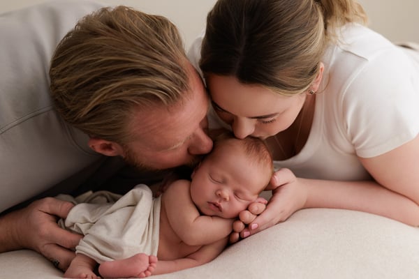 Two parents kissing above their sleeping newborn, who lies wrapped between them