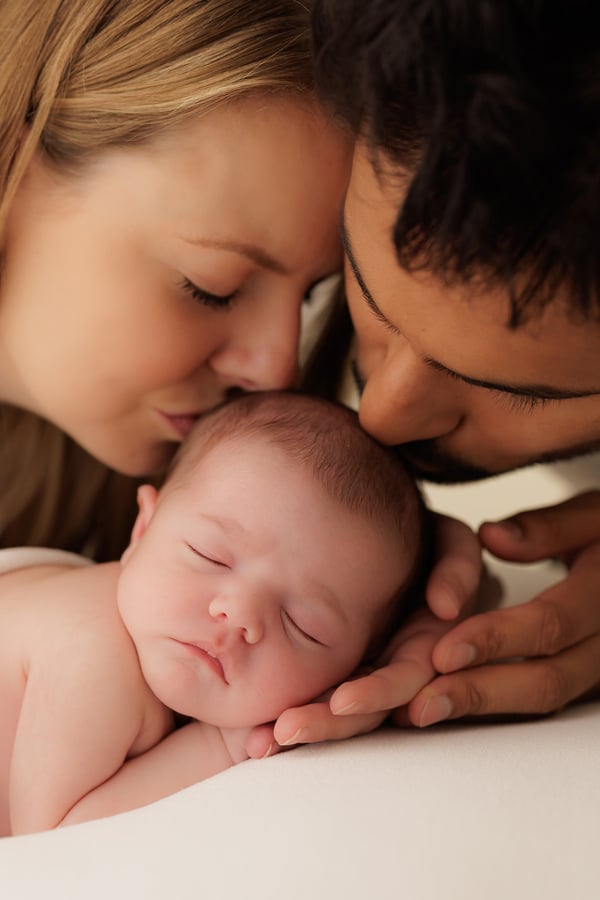 Both parents leaning in together to kiss their sleeping newborn