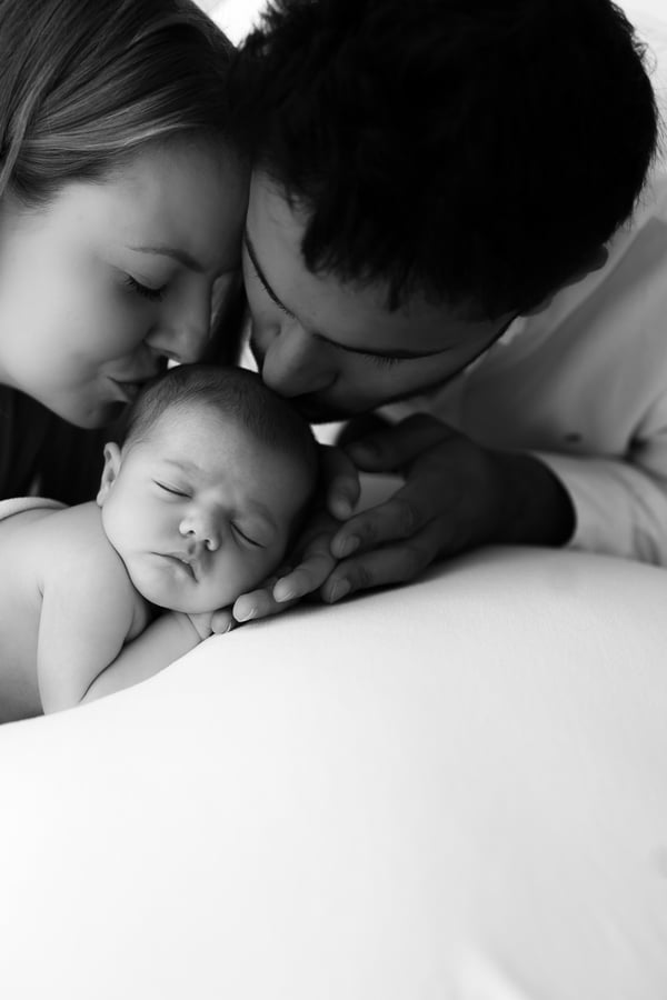 Both parents leaning in to kiss their sleeping newborn's head, in black and white