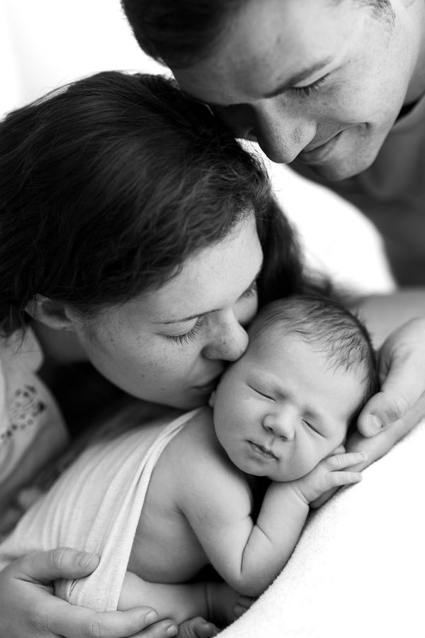 Both parents leaning over their swaddled newborn, the mother kissing the baby's head, in black and white