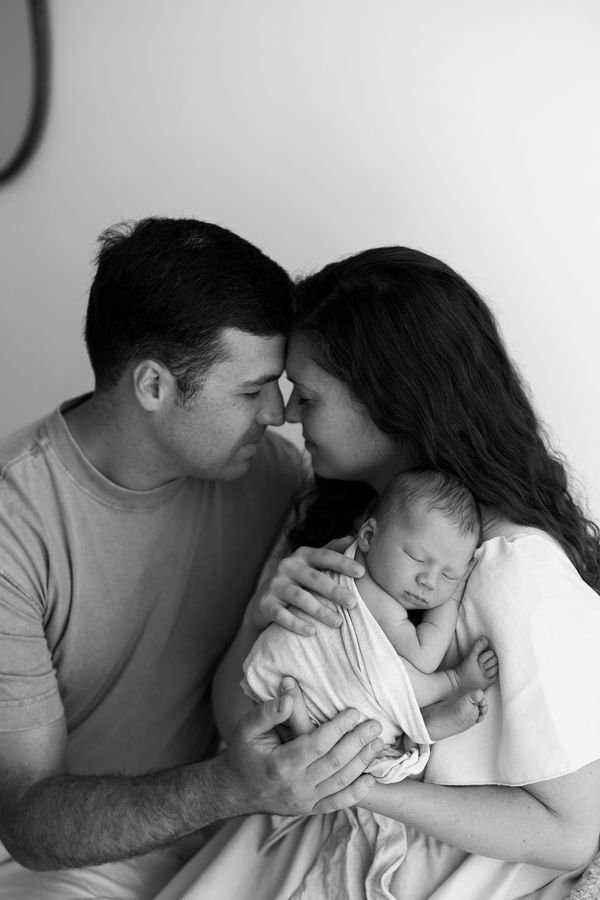 Both parents nose to nose while holding their swaddled newborn, in black and white