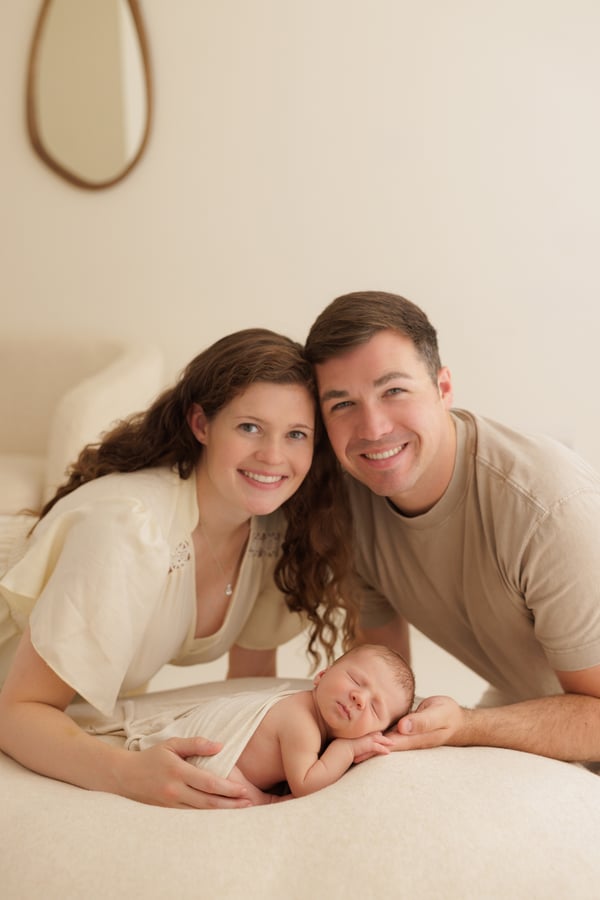 Both parents lying down with their sleeping newborn, smiling at the camera