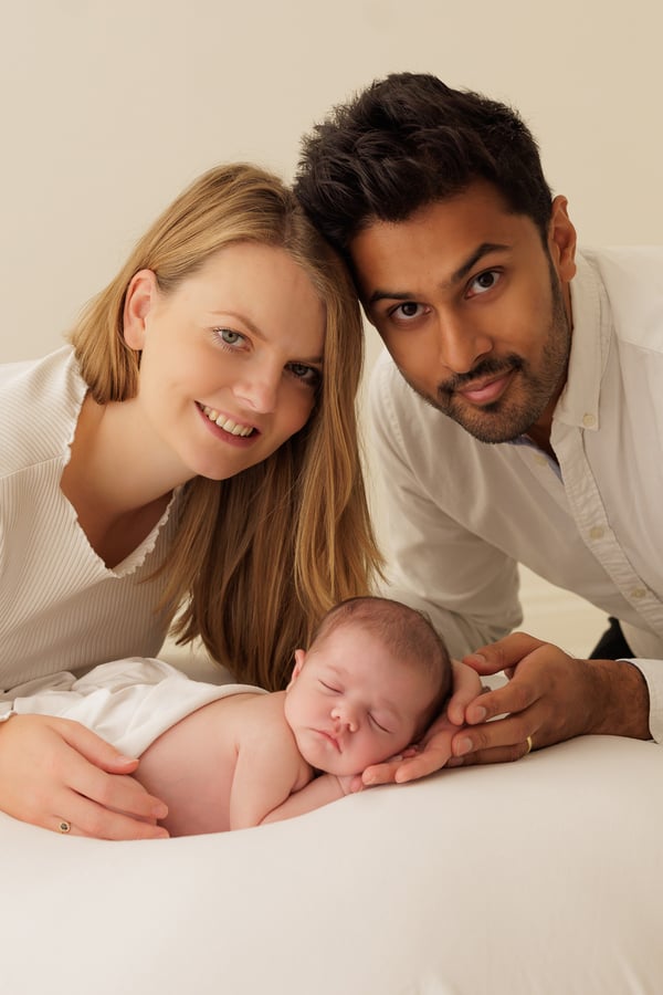 Both parents smiling at the camera from behind their sleeping newborn