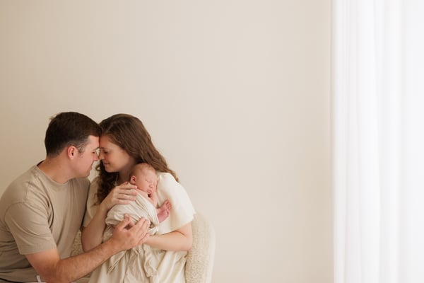 Both parents standing by a window holding their newborn, the studio wall bare around them